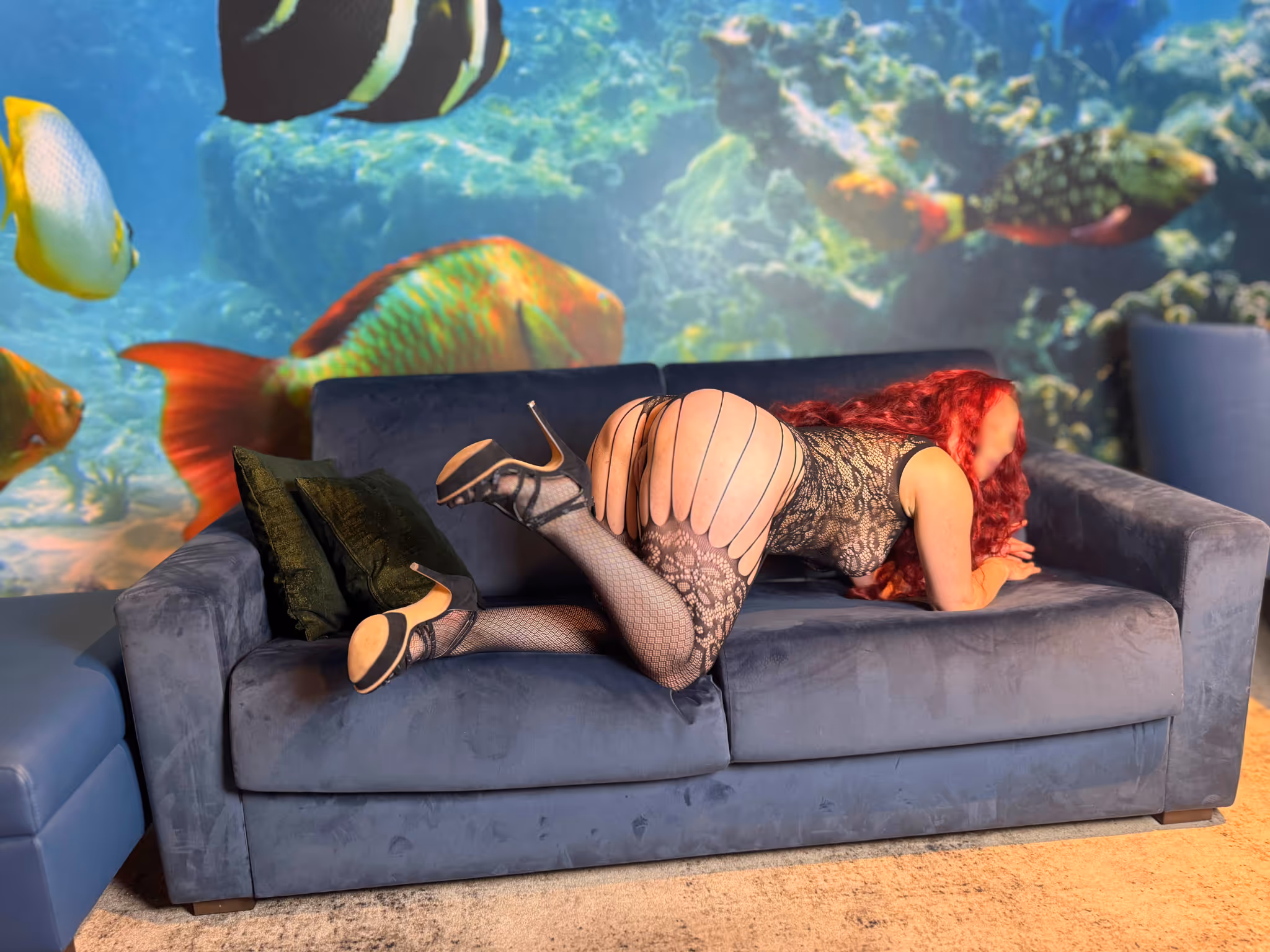 Escort Romy Bondage in Dresden kniet gefesselt auf dem blauen Samt Sofa. Lange rote Locken fallen über den Rücken Hände auf dem Rücken gebunden schwarze Netzstrümpfe mit Strapsen High Heels sinnliche und unterwürfige Bondage Pose vor Aquarium Wandbild Luxus Hotel Dresden.